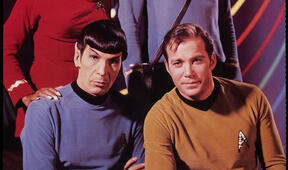 Spock and Kirk from Star Trek posing together for a group photo, sitting. Behind them, Uhura and Bones are standing, cropped out of the picture.